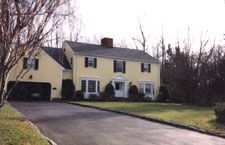 A large yellow house with black shutters and a driveway leading to it.