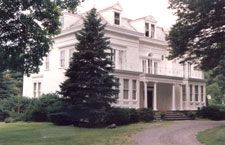 A large white house with a driveway leading to it is surrounded by trees and bushes.