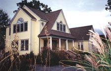 A large yellow house with a porch is surrounded by tall grass.