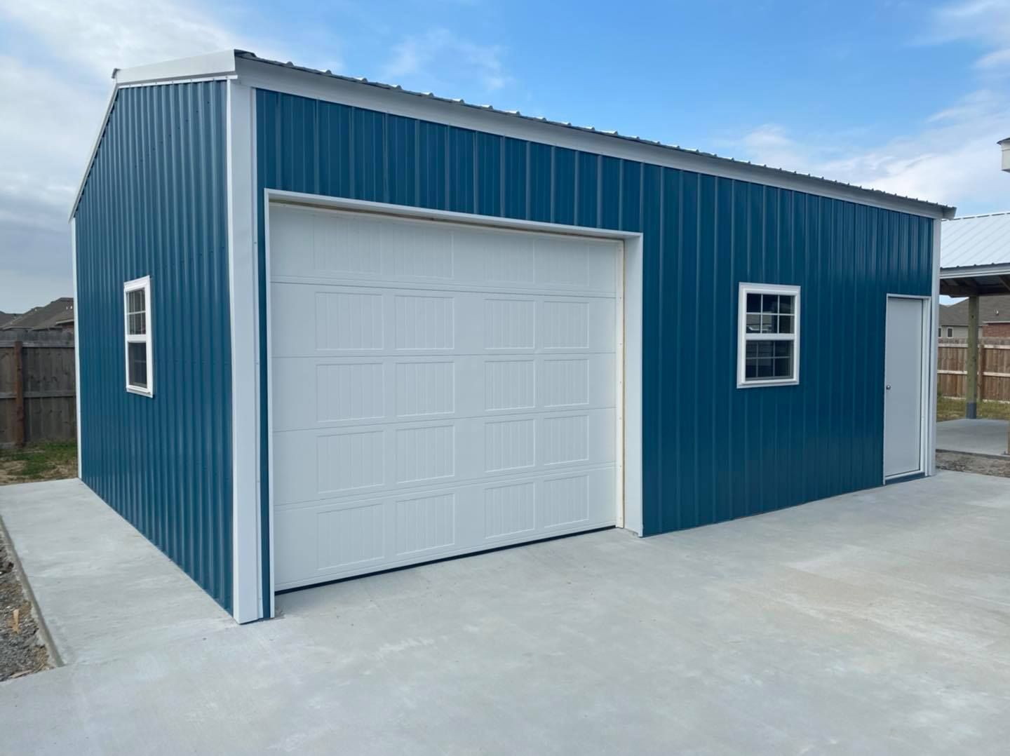 Blue metal garage with white door and trim, two windows, and concrete pad.