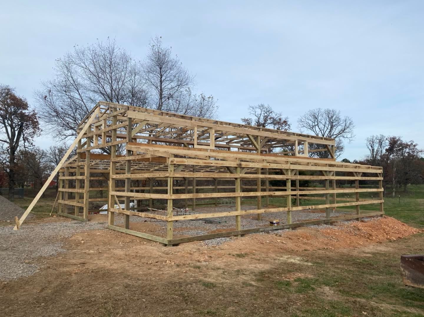 Wooden barn frame under construction on a gravel surface, trees in the background.