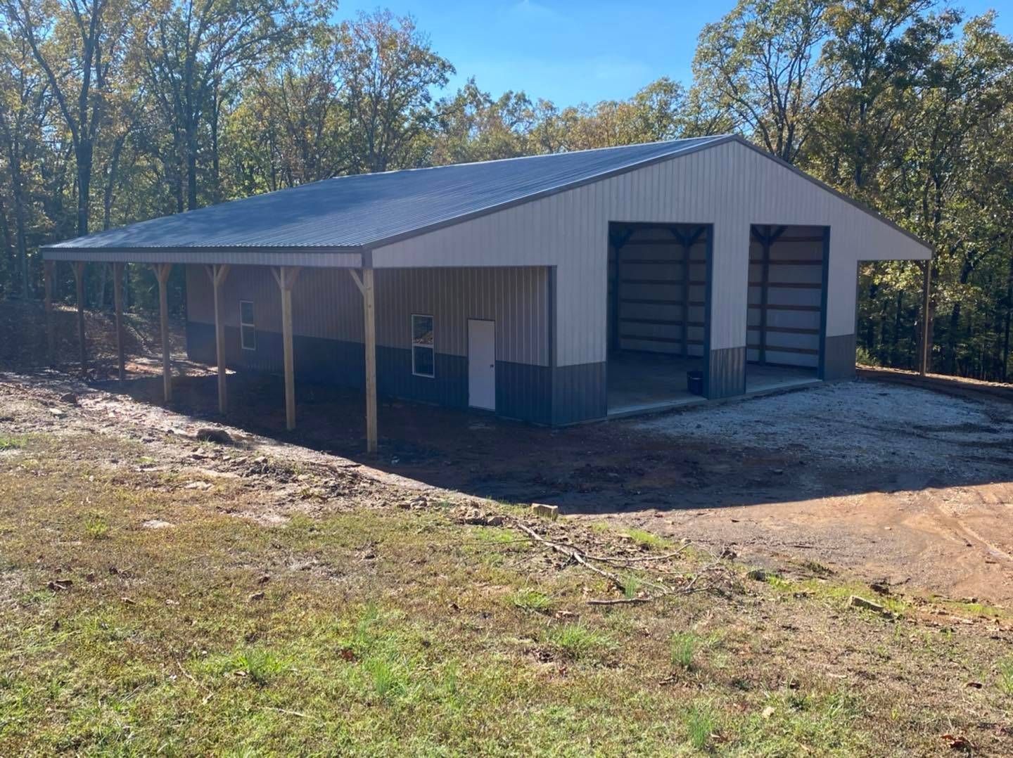 Large metal building with a two-bay garage, carport, and side door. The structure is in a wooded area.