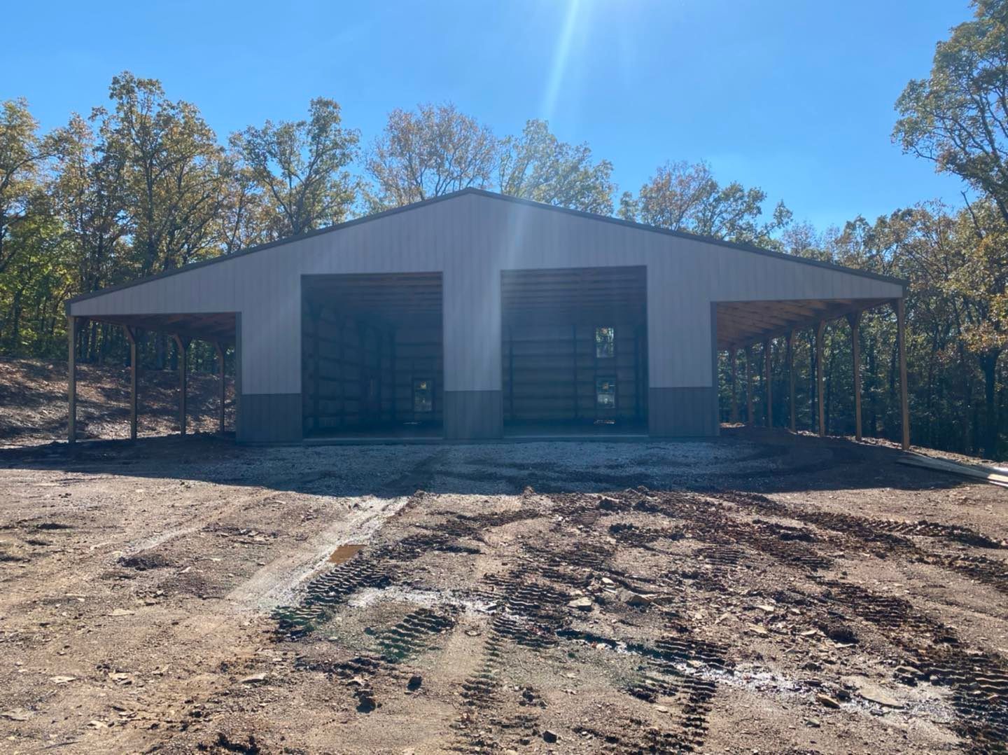 Metal building with two garage doors and canopies, on a dirt clearing, trees in background.