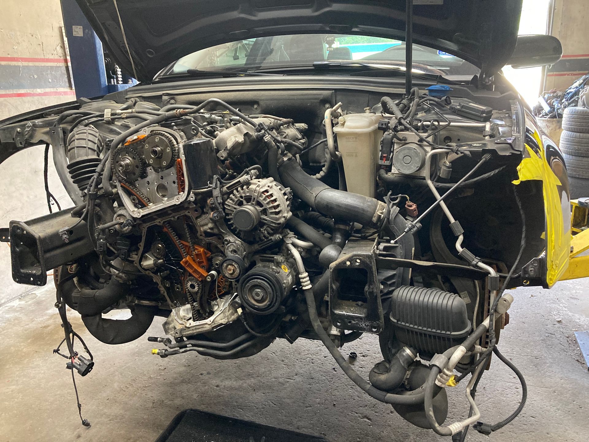 Partially disassembled car engine bay with exposed components in a workshop, hood open.