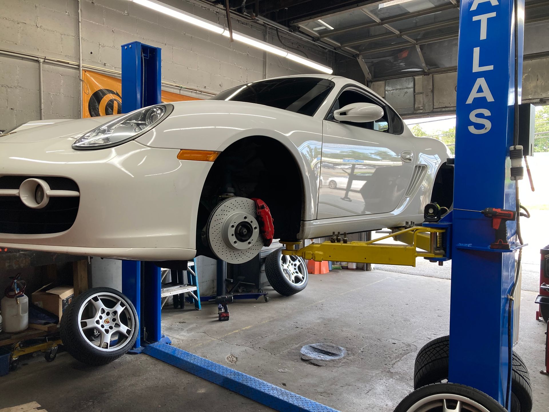 White sports car raised on a blue lift in an auto repair shop, front wheel removed exposing the brake rotor.