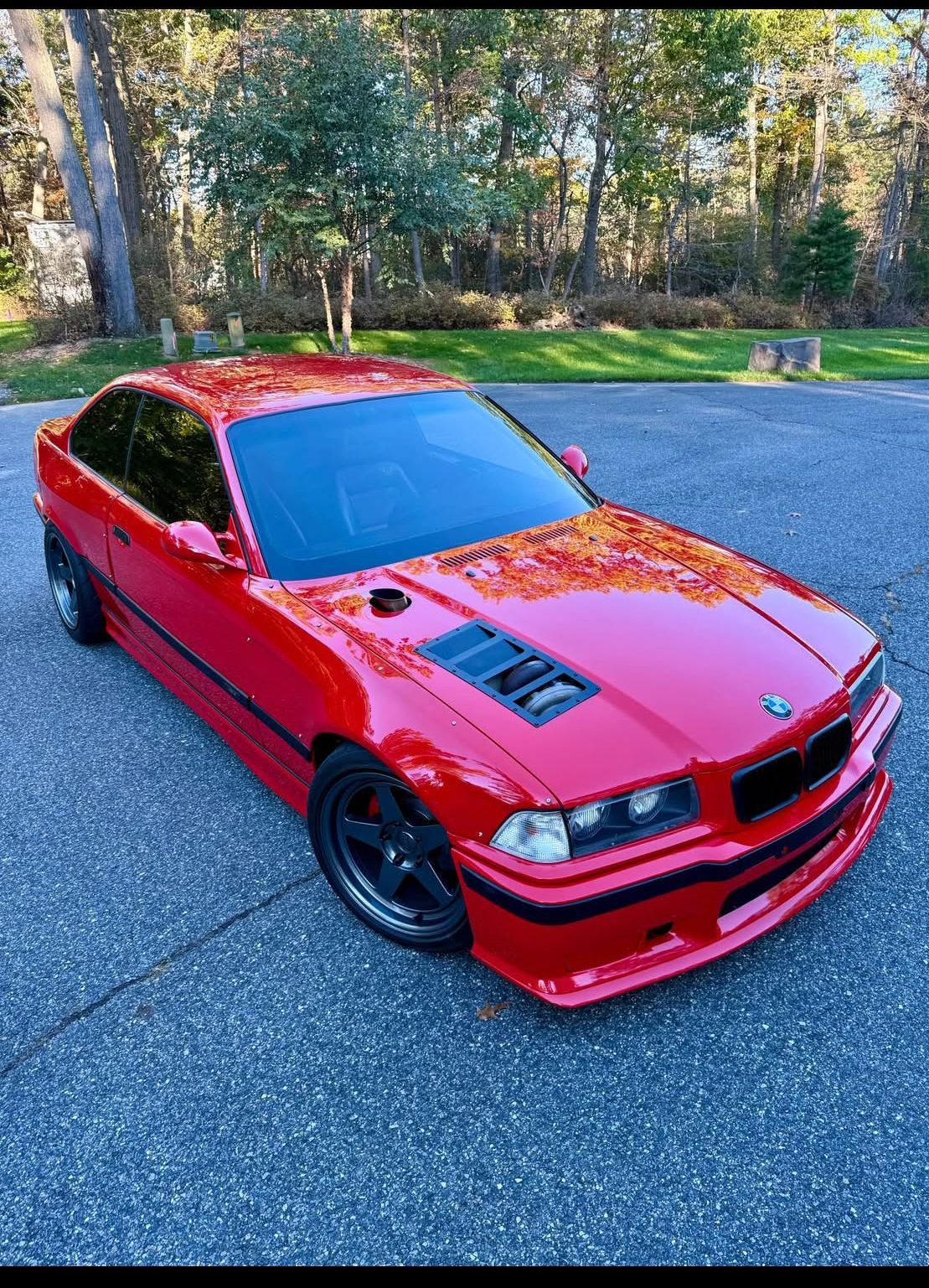 Red sports coupe with black wheels parked on a paved road beside trees and grass.