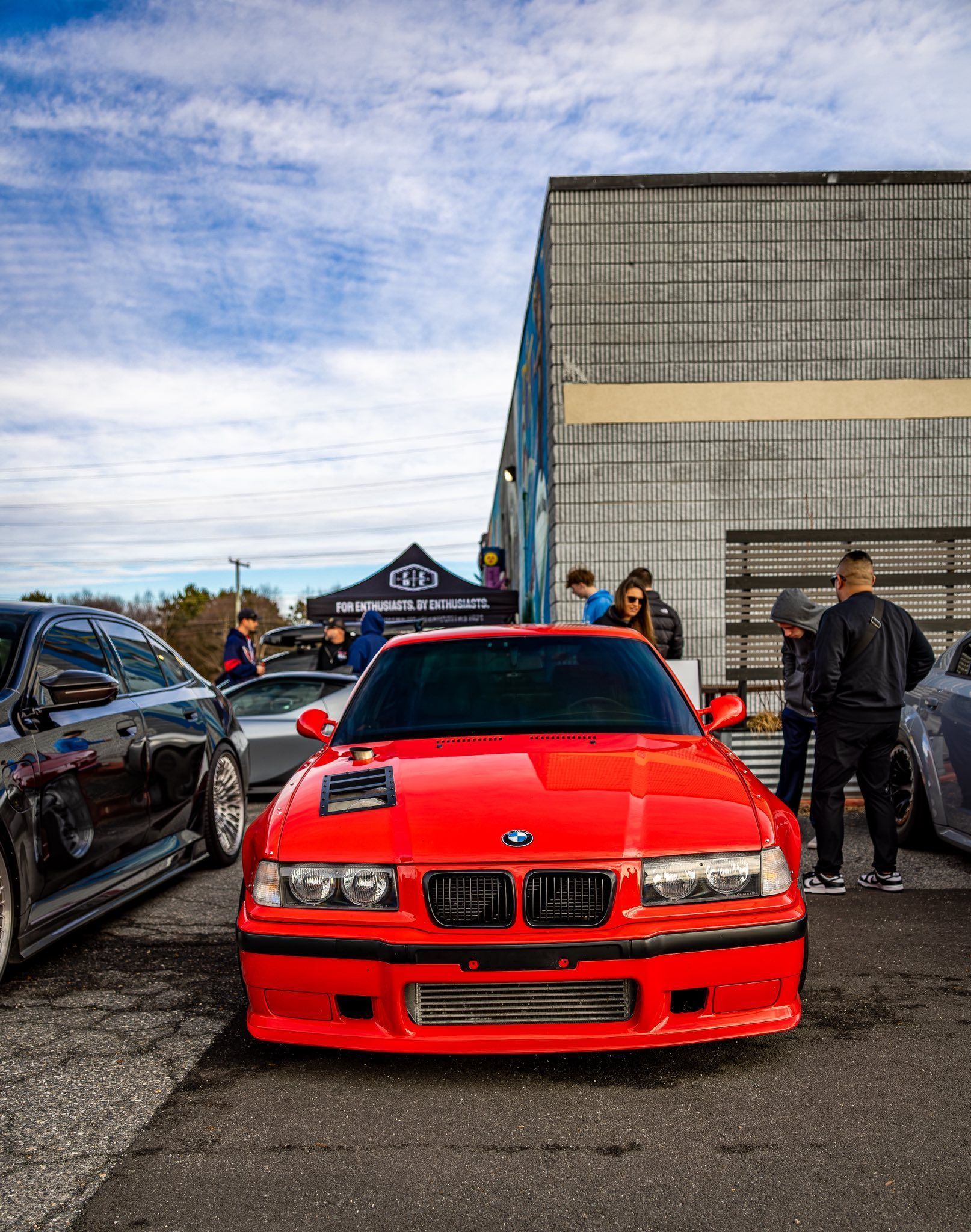 Red sports car parked at a car meet beside a gray building, with people standing nearby.