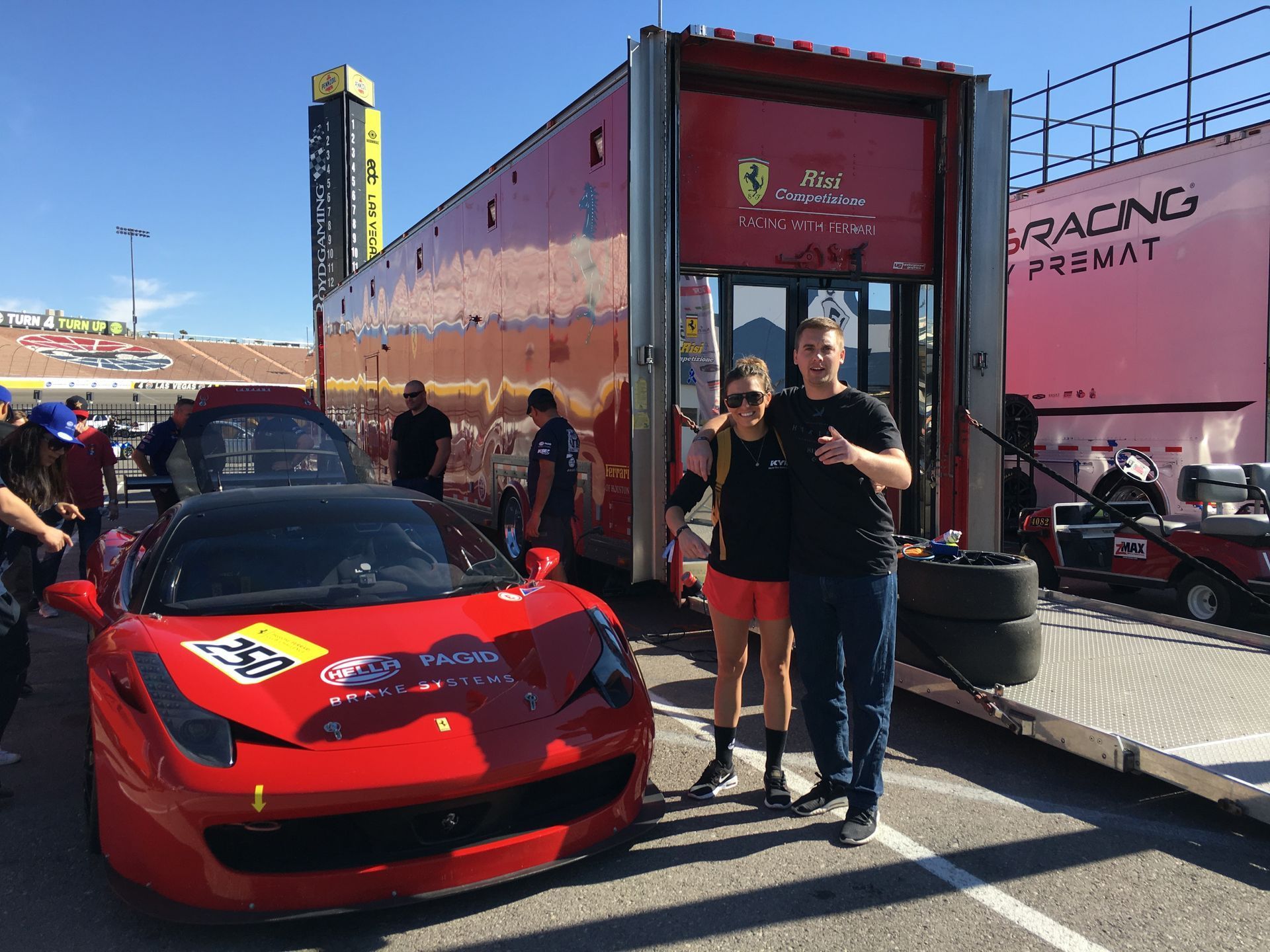 Red race car in front of a trailer, with two people posing beside it at a racetrack pit area.