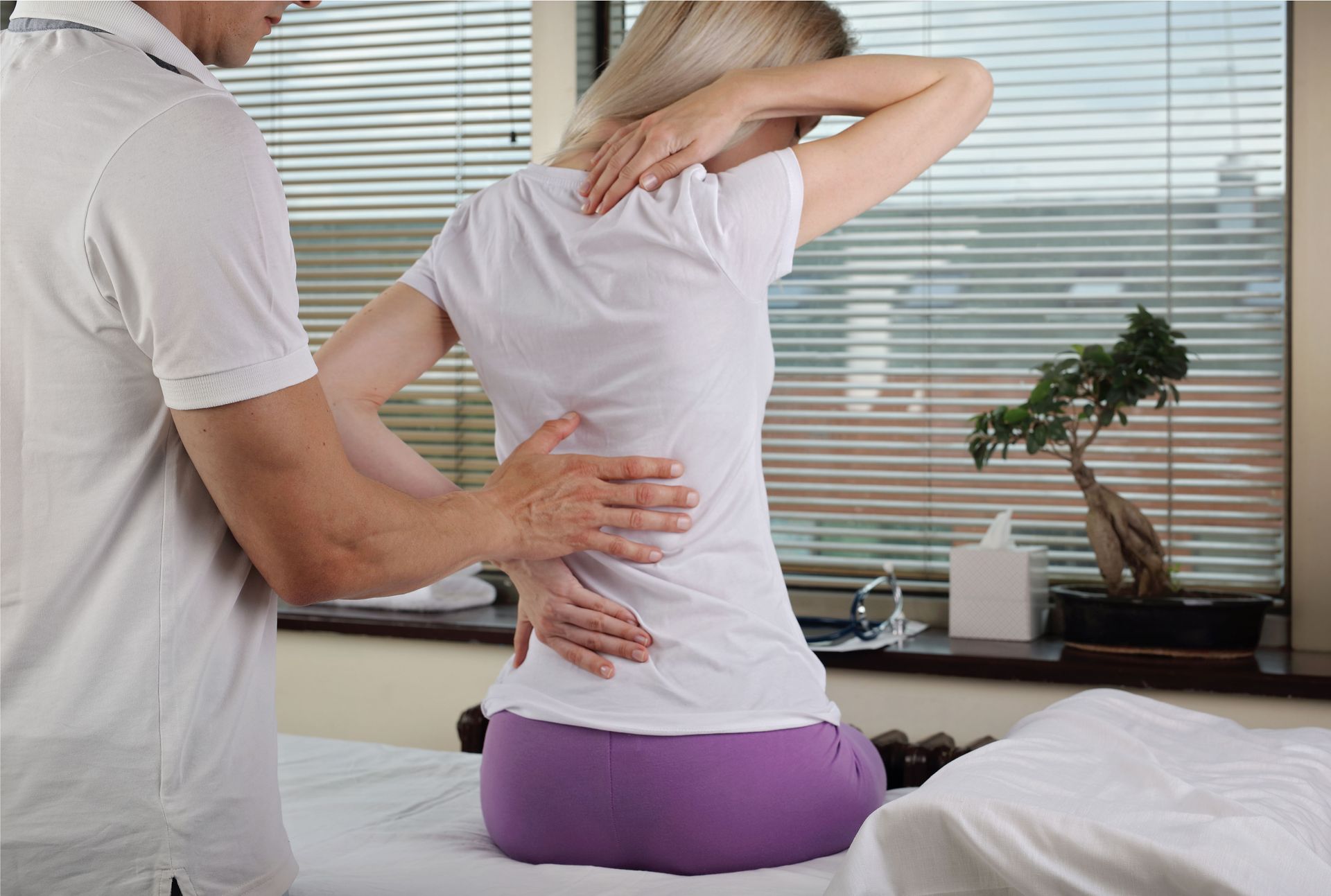 Therapist examines woman's back, seated, in a clinic with a bonsai tree and a window. She holds her neck in pain.