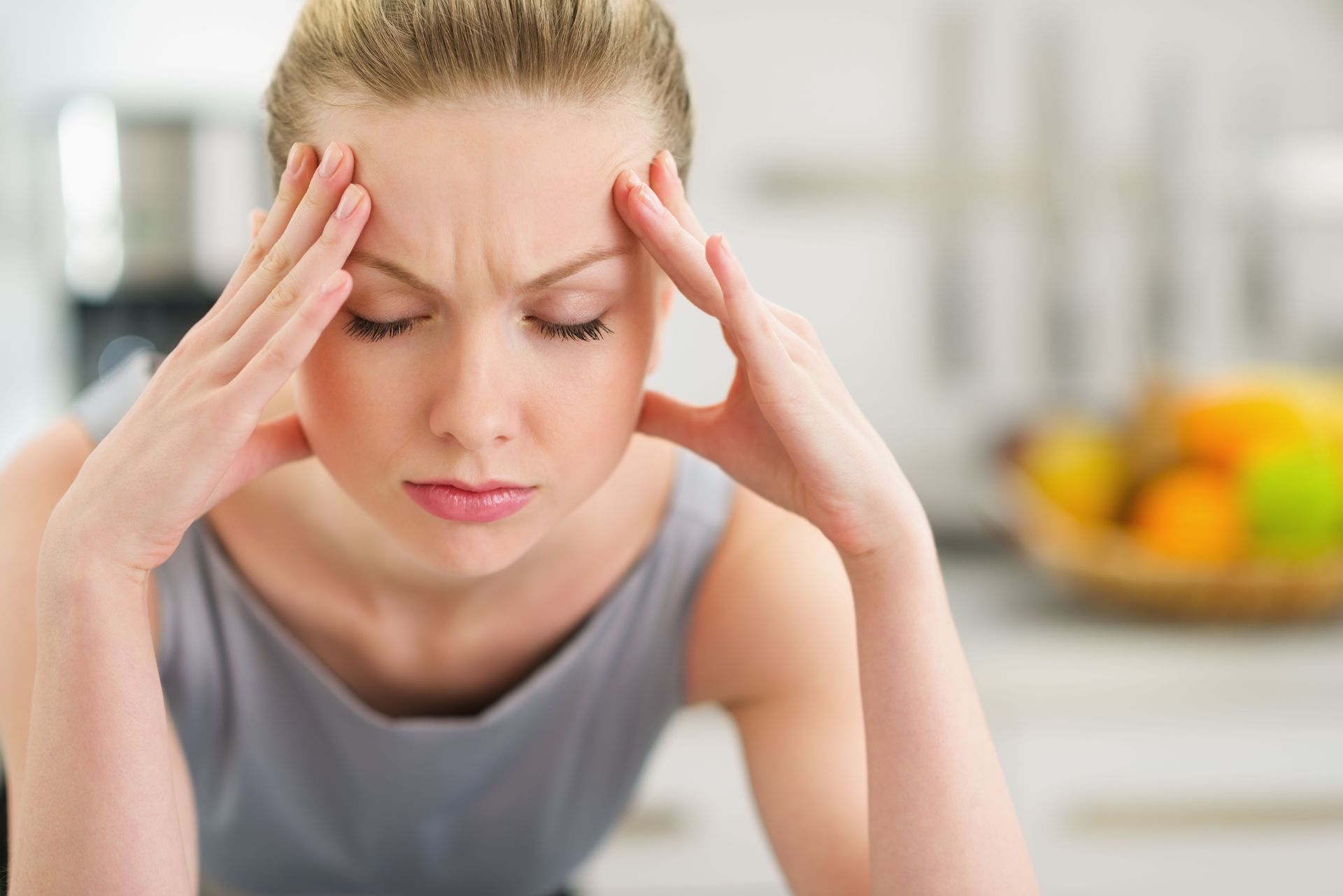 Woman with headache, holding temples in kitchen, eyes closed, concerned expression.