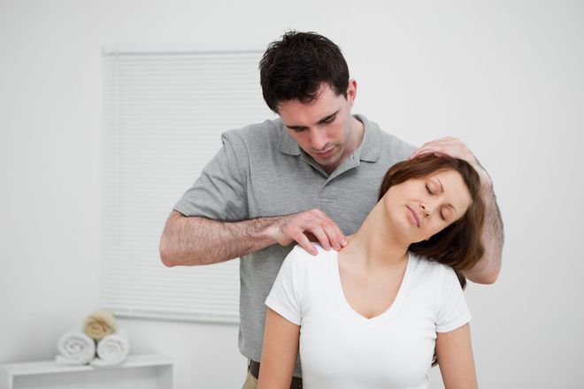 A man gently stretches a woman's neck in a bright room; they are indoors, likely at a clinic.