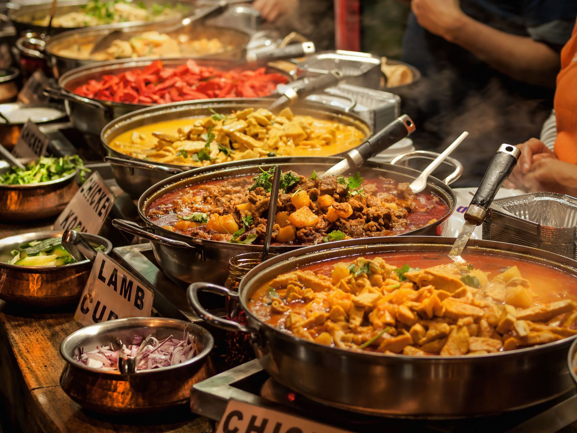 A buffet-style counter features several metal pots filled with various curries, meats, and sliced red onions.