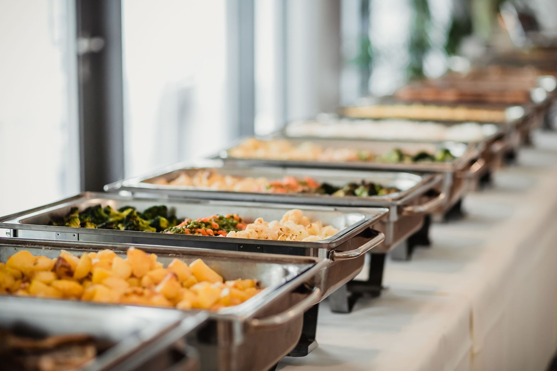 A long buffet table featuring multiple metal chafing dishes filled with warm food, including roasted potatoes and greens.