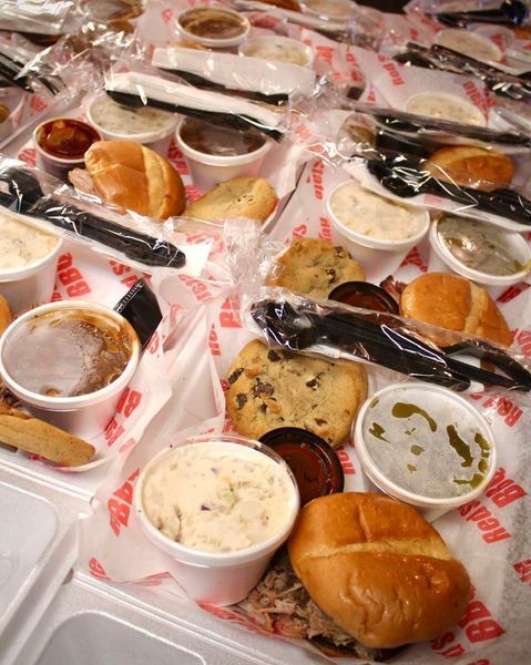 Overhead view of several takeout containers filled with BBQ sandwiches, sides, cookies, and sauce cups on printed paper.