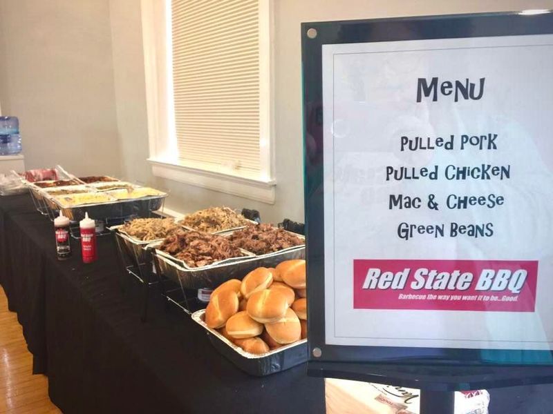 A Red State BBQ catering buffet table featuring trays of food, buns, and a menu board listing meats and side dishes.