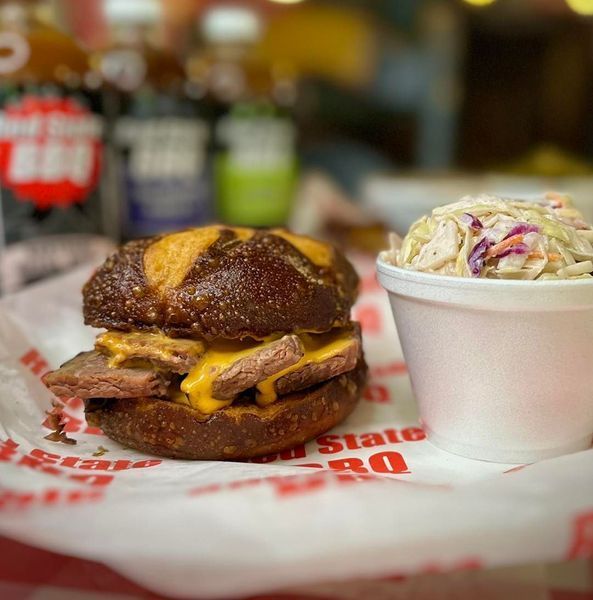 A brisket sandwich on a pretzel bun with melted cheese, served with a side of coleslaw on paper in a restaurant.