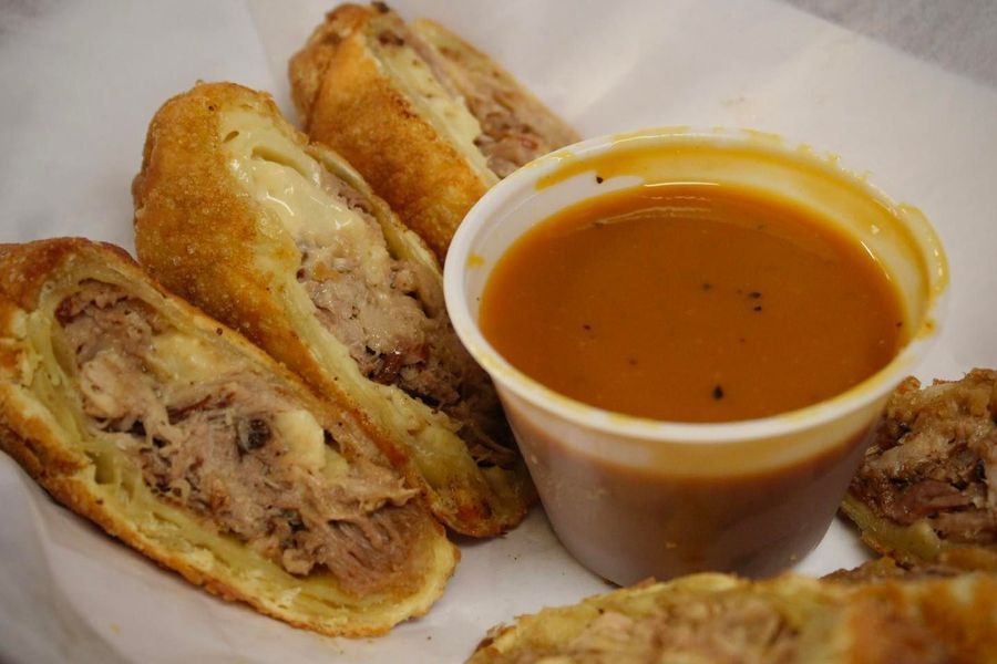 Deep-fried, meat-filled egg rolls halved on white paper next to a plastic cup of brown dipping sauce.