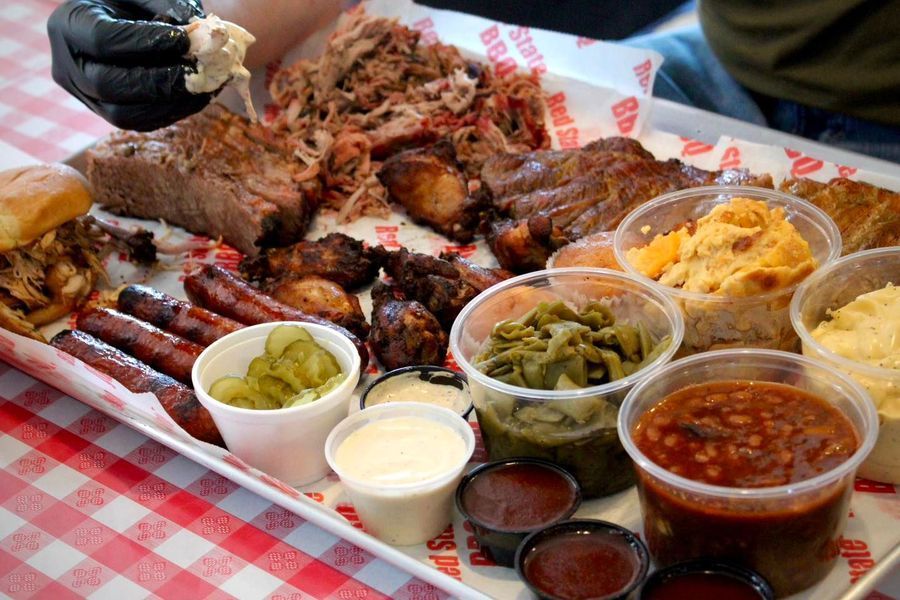A large metal tray filled with various smoked meats, sausages, sides, and sauces on a red and white checkered tablecloth.
