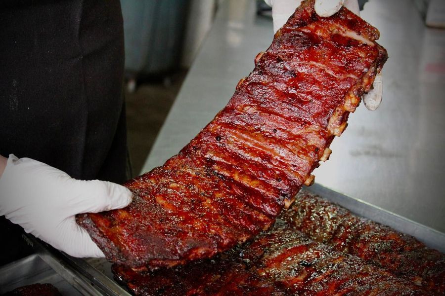 A pair of gloved hands holds a rack of glazed, cooked ribs above a metal tray of more ribs.
