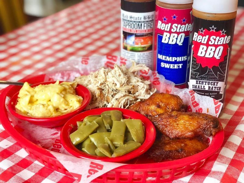 A barbecue platter with chicken, green beans, and mac and cheese in a red basket, served with three bottles of BBQ sauce.