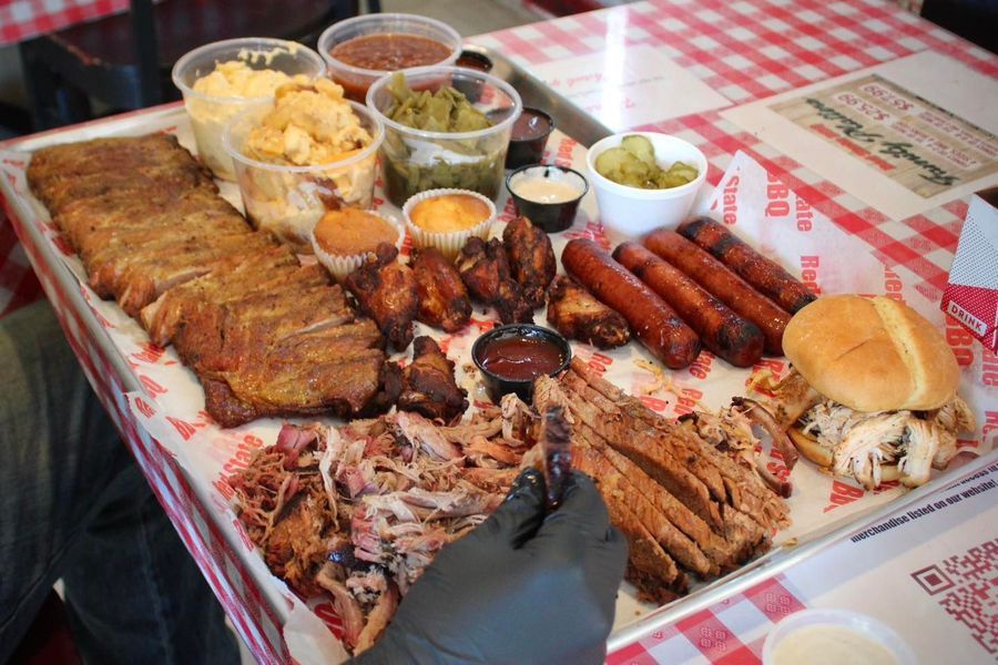 A large metal tray piled with BBQ ribs, pulled pork, brisket, sausages, wings, sides, and a sandwich on a checkered table.