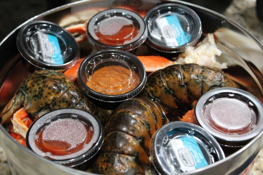 A silver metal container filled with cooked lobster tails, crab legs, and various small cups of dipping sauces.