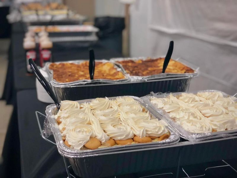 Aluminum catering pans on a buffet table, featuring banana pudding with whipped cream and baked pasta dishes.