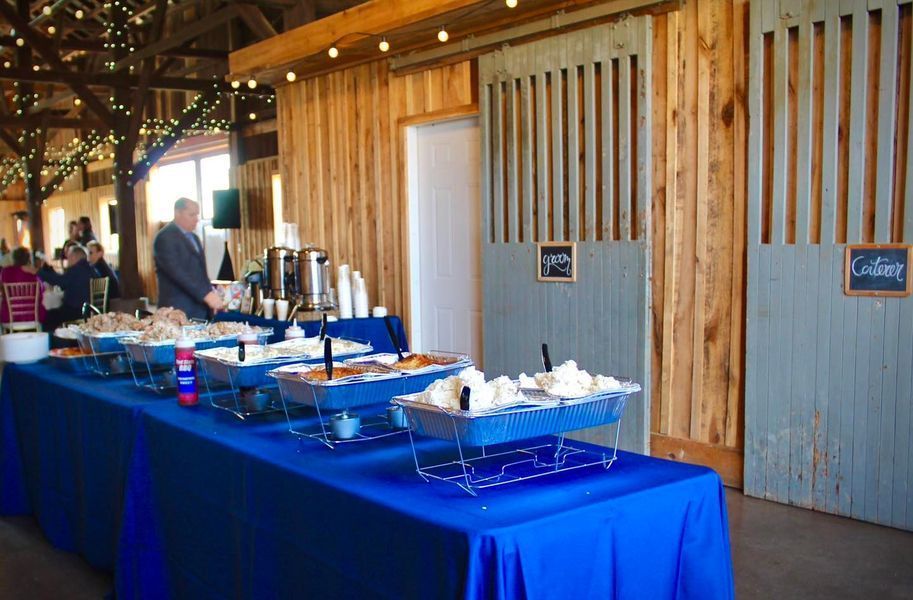 A buffet line of chafing dishes on a blue-covered table inside a rustic wooden barn with string lighting overhead.