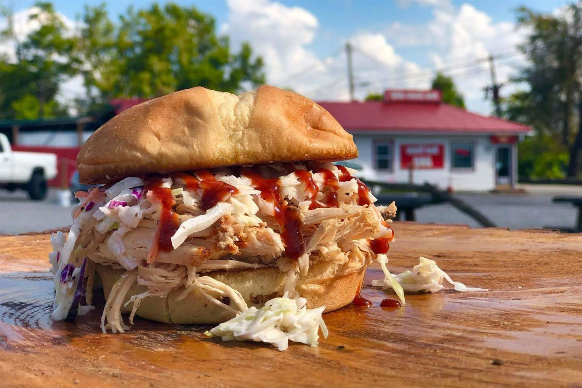 A pulled pork sandwich with coleslaw and BBQ sauce on a wooden surface, with a blurred roadside restaurant in the back.
