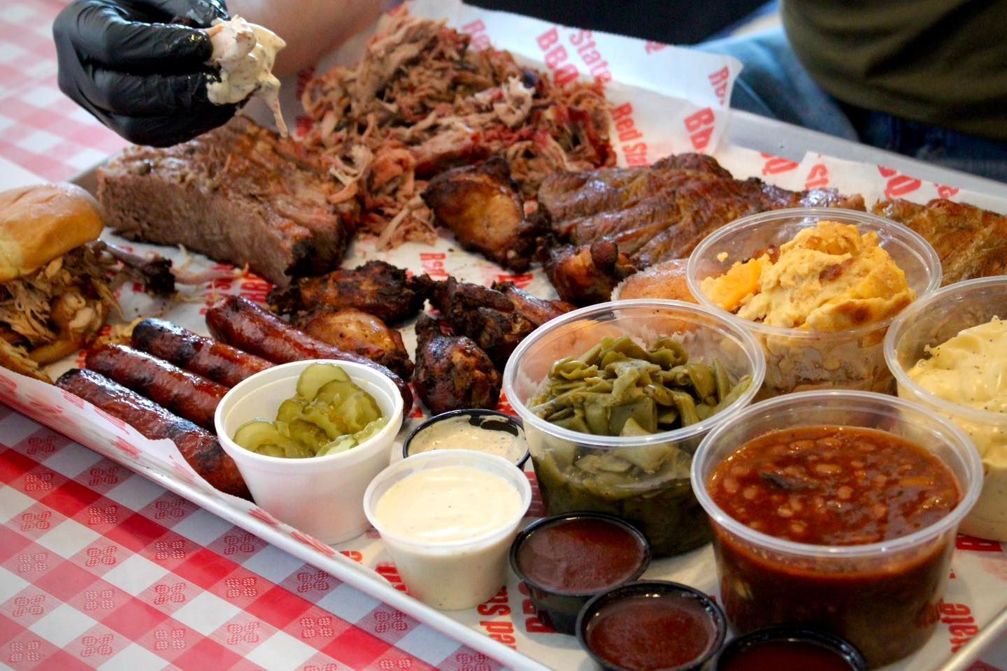 A platter of barbecue meats, sides, and sauces on a red and white checkered tablecloth, with a gloved hand picking meat.