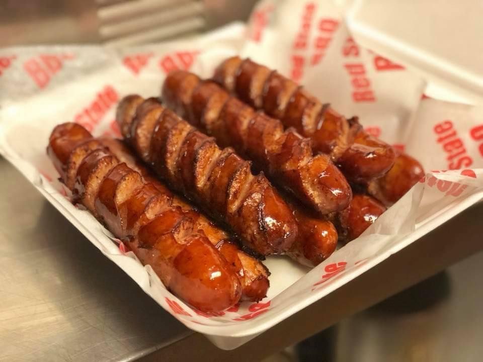 Four grilled sausages with diagonal scores, coated in a dark brown glaze, served on parchment paper in a white tray.