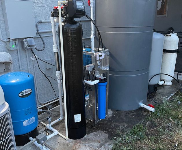 Outdoor water treatment equipment with blue tank, black filter, and gray storage tank beside a house