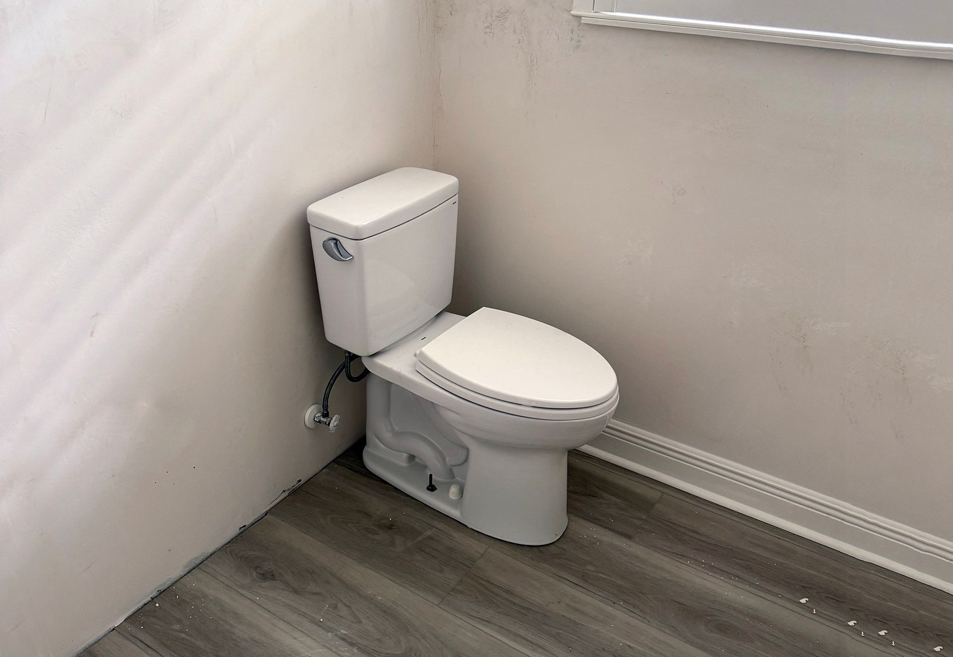 Small white toilet in a bathroom corner with gray wood-look flooring and white walls.