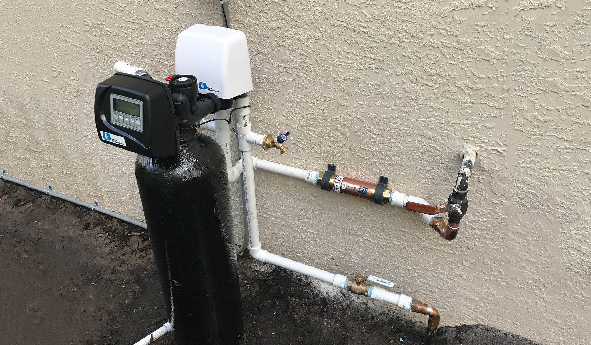 Water meter and exposed piping mounted on a beige exterior wall.
