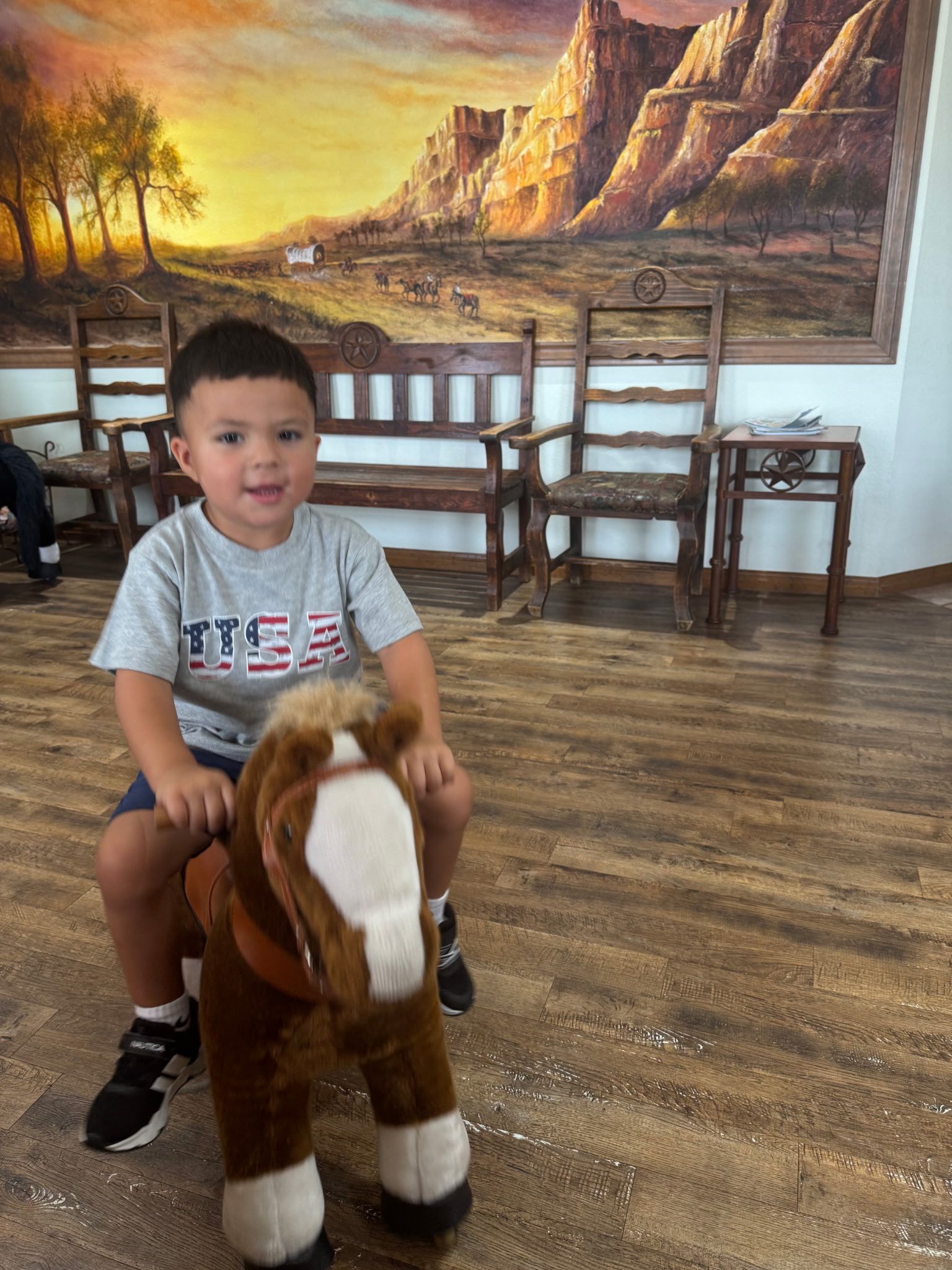 Child sitting on a brown toy horse in a room with wooden chairs and a large landscape mural