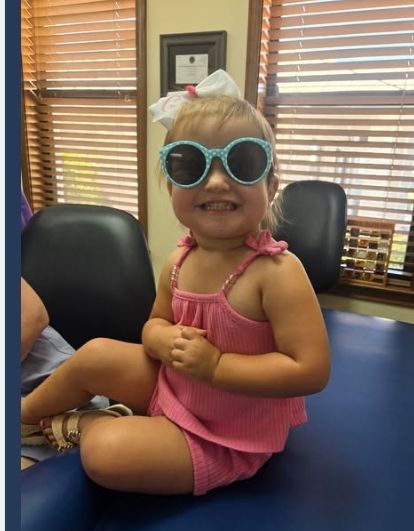 Smiling child in pink outfit and blue sunglasses sitting on a chair in a waiting room