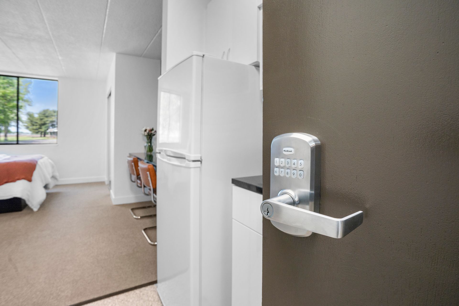 A door with a keypad lock is ajar, revealing a room with a bed, refrigerator, and chairs.