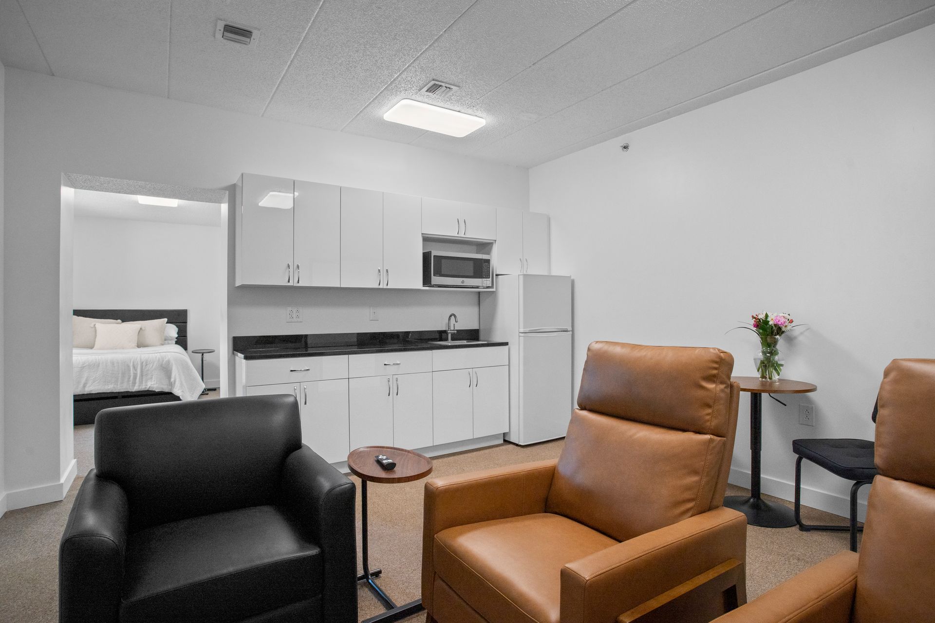 A bright apartment with a small kitchen, two recliners, and a bedroom visible in the doorway.