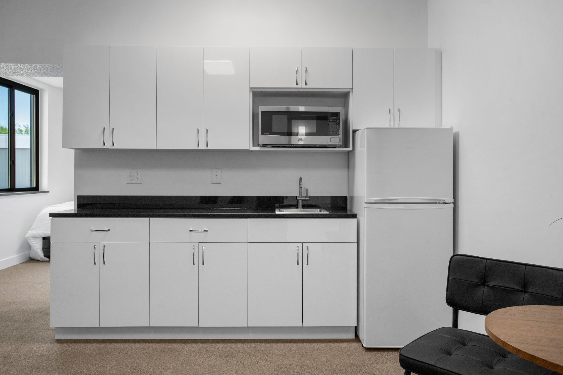 White kitchen cabinets, microwave, refrigerator, and small table with a black chair.