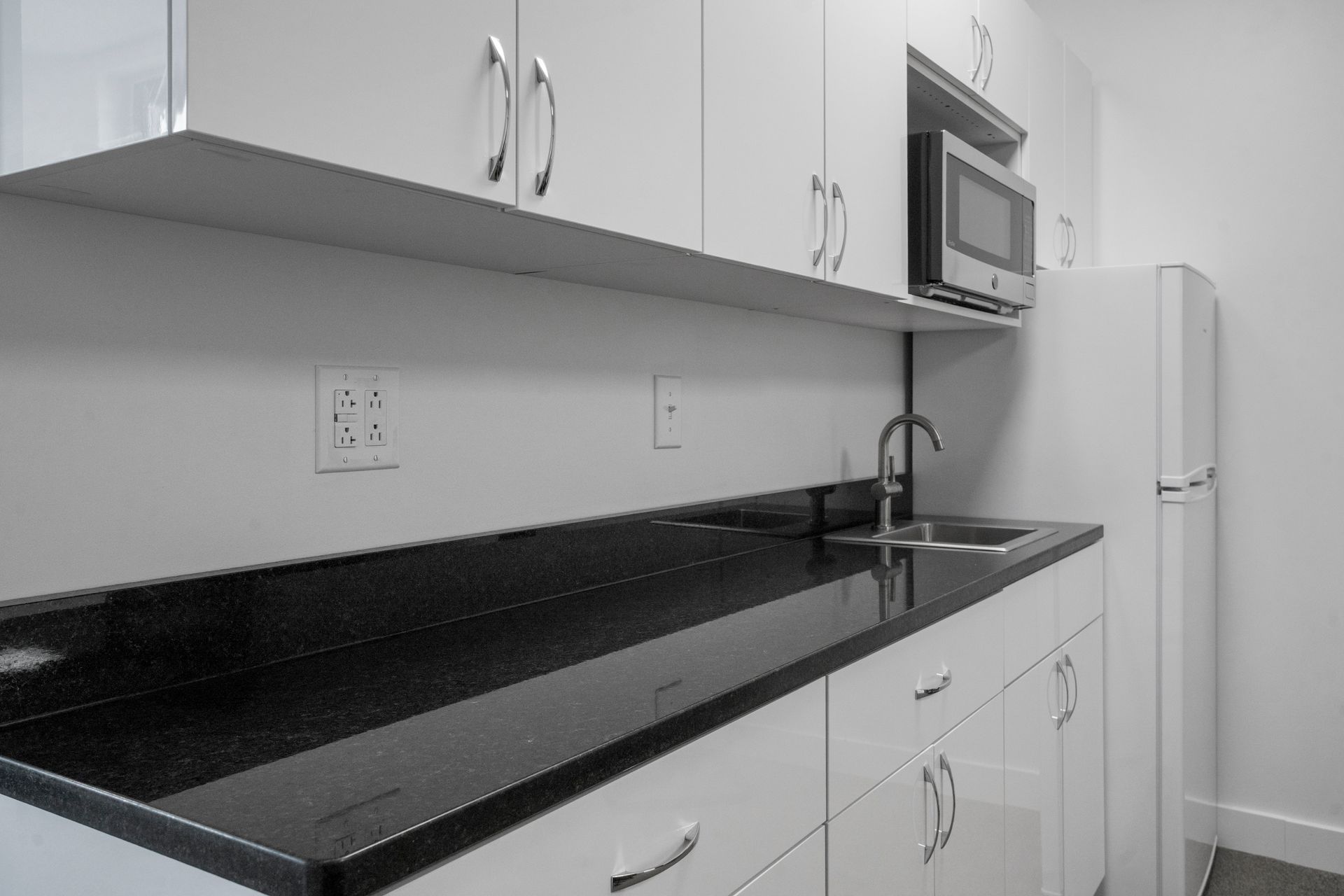 White kitchen with dark countertop, cabinets, sink, microwave, and refrigerator.