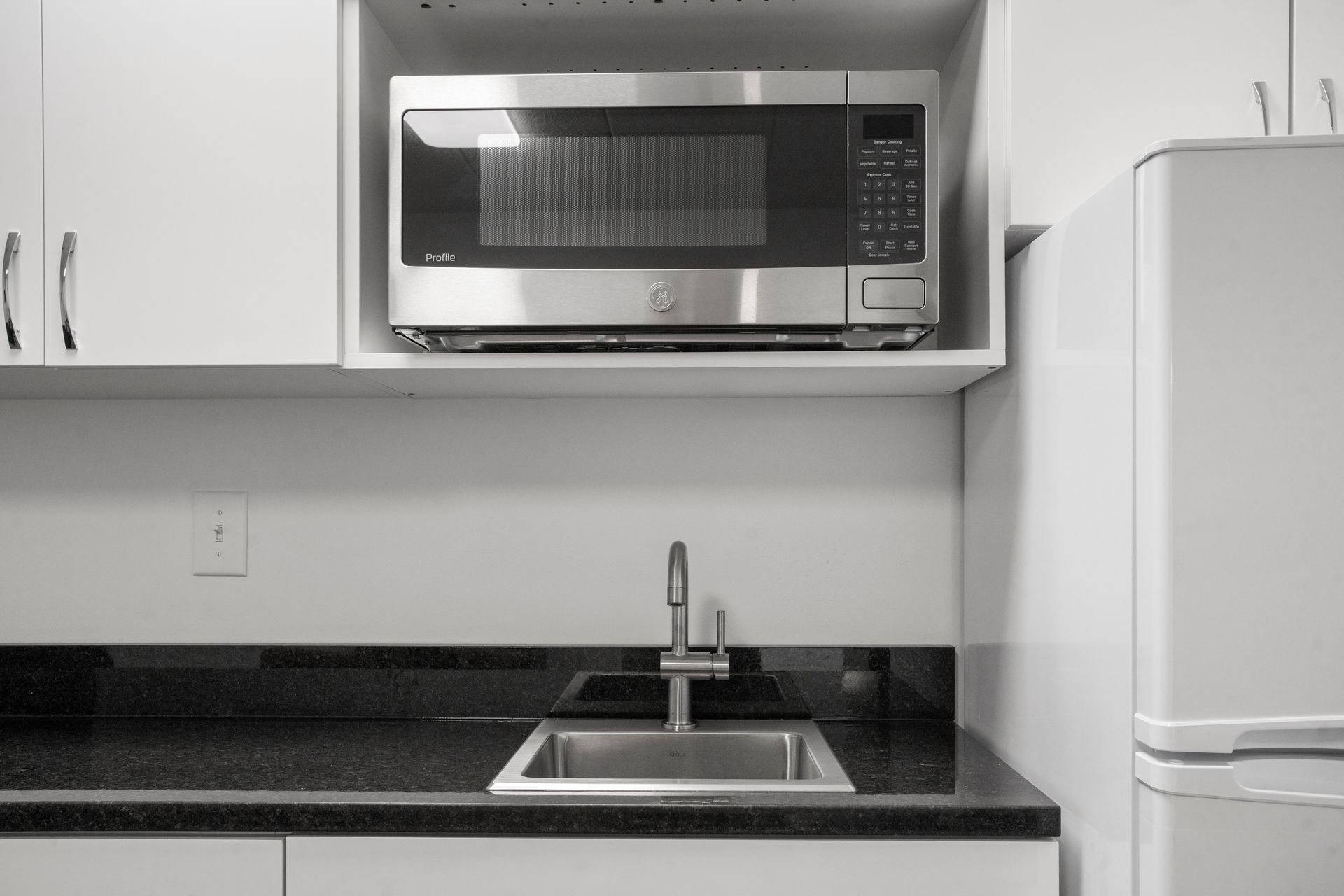 Kitchen with microwave, sink, and refrigerator. White cabinets and black countertop.