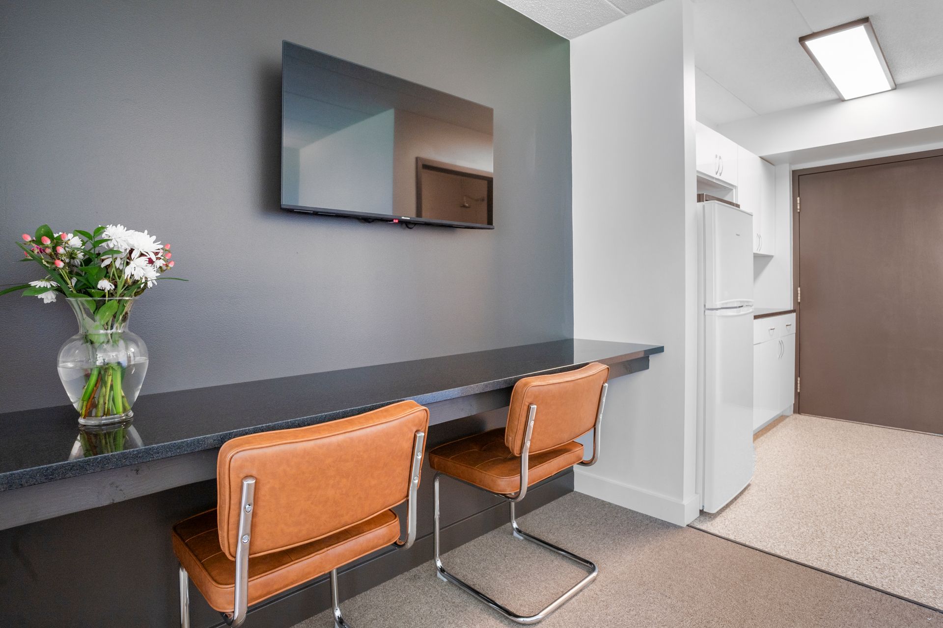 A small office nook with a table, two brown chairs, a TV, flowers, and a mini-kitchen.
