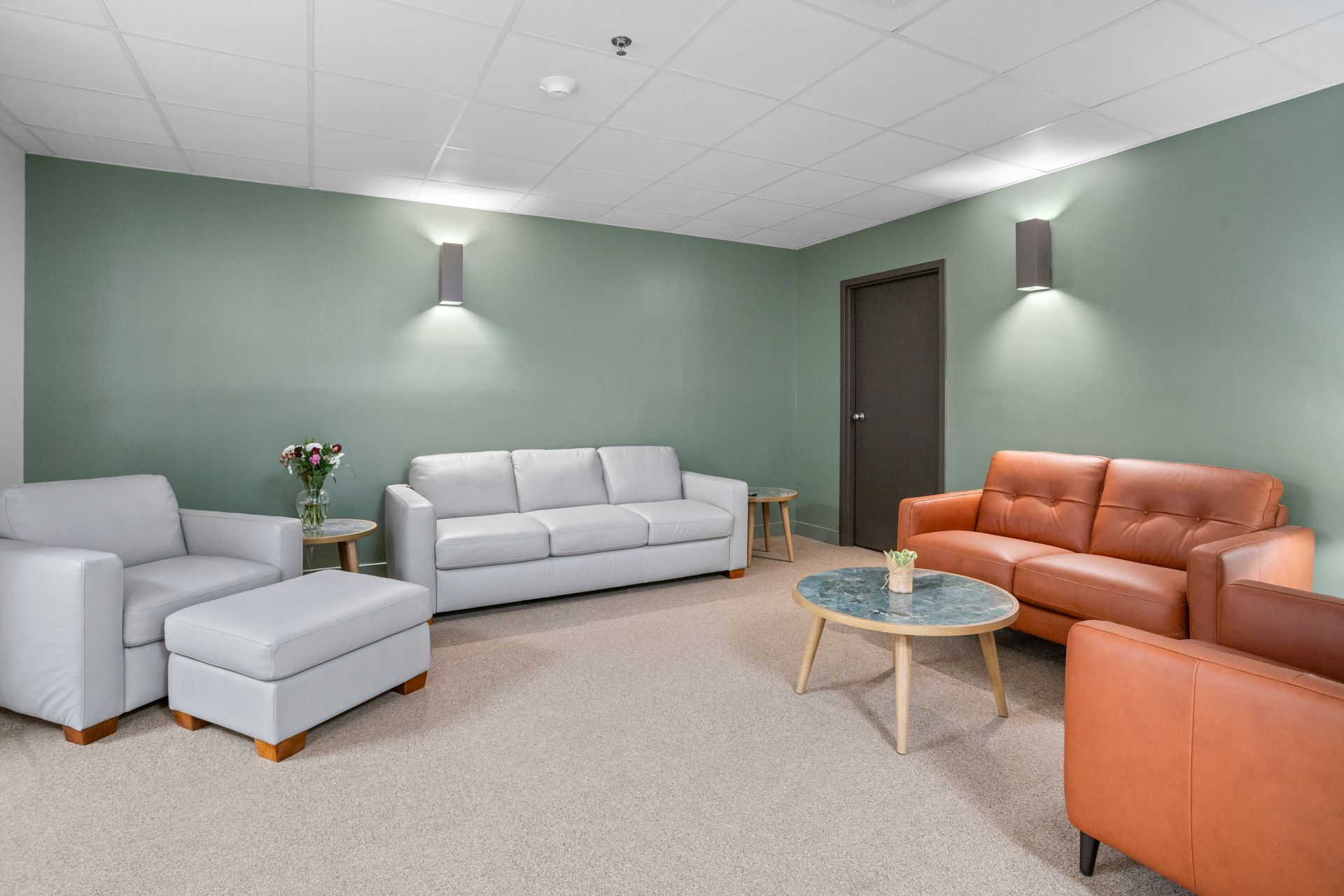 A waiting room with two sofas, armchairs, coffee table, and a door. Green and tan color scheme.