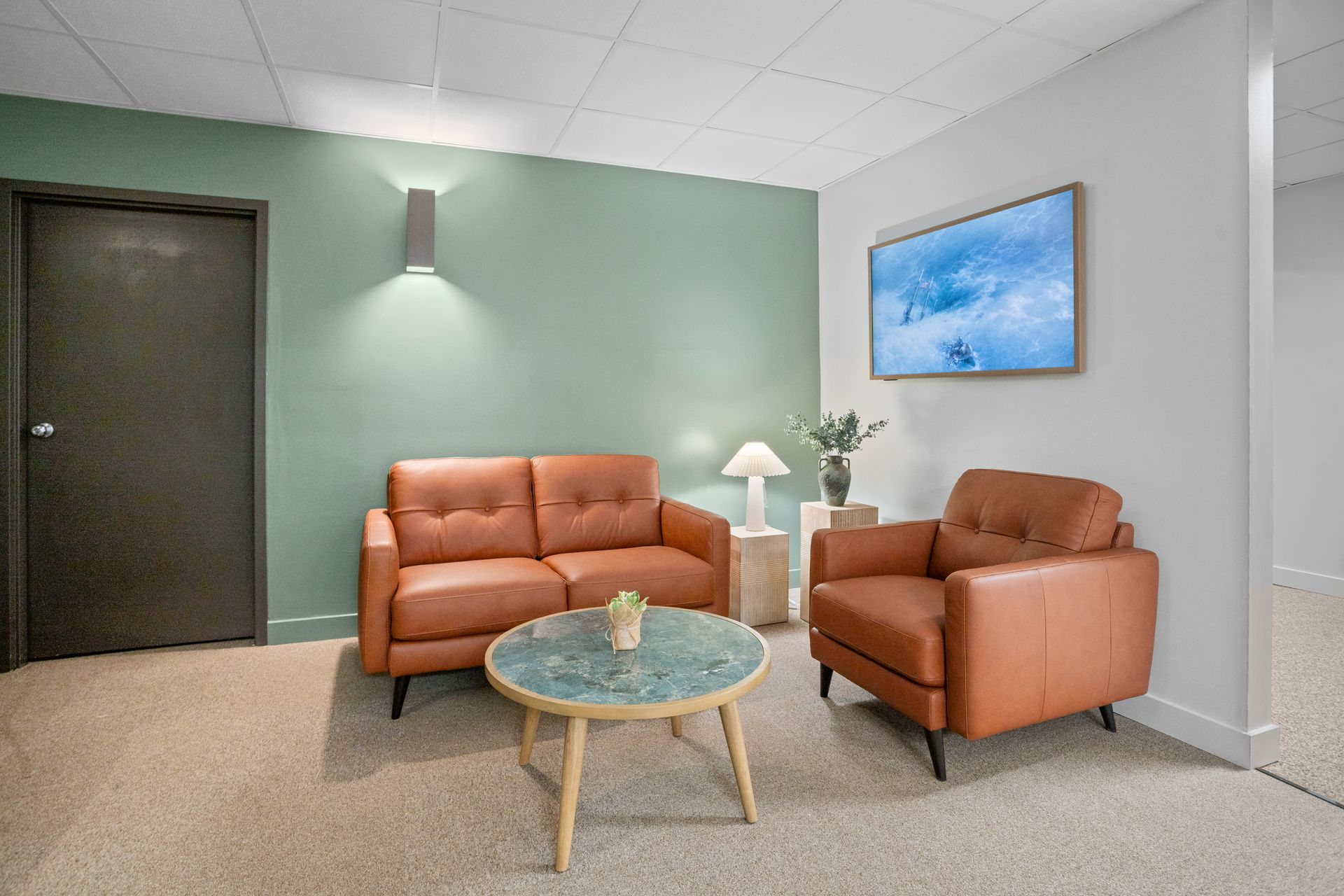 Waiting area: brown leather couch and chair, light green wall, TV, coffee table, tan carpet.