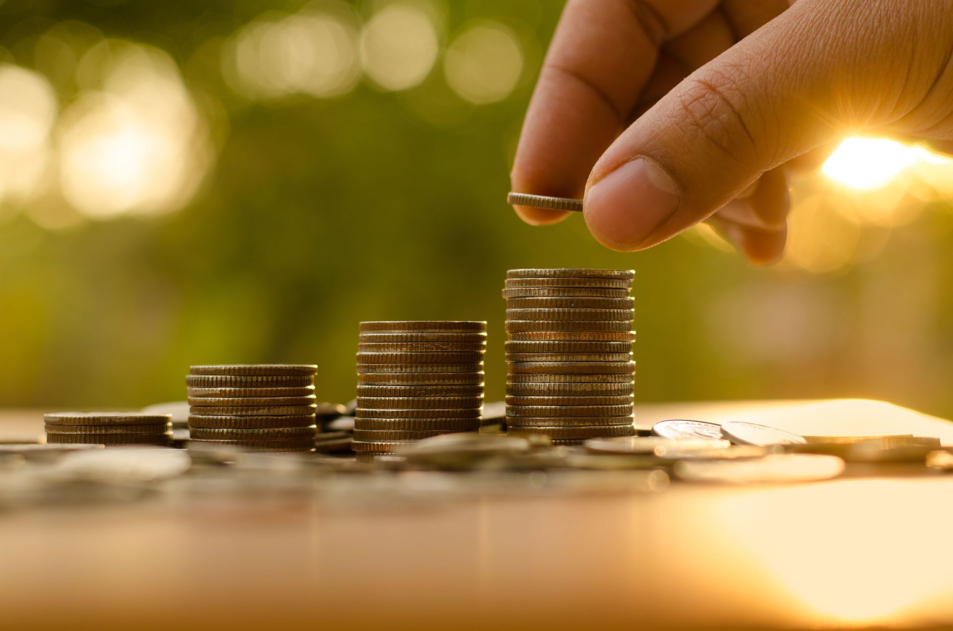 Hand placing a coin on a stack of coins, growing a money pile in an outdoor setting.
