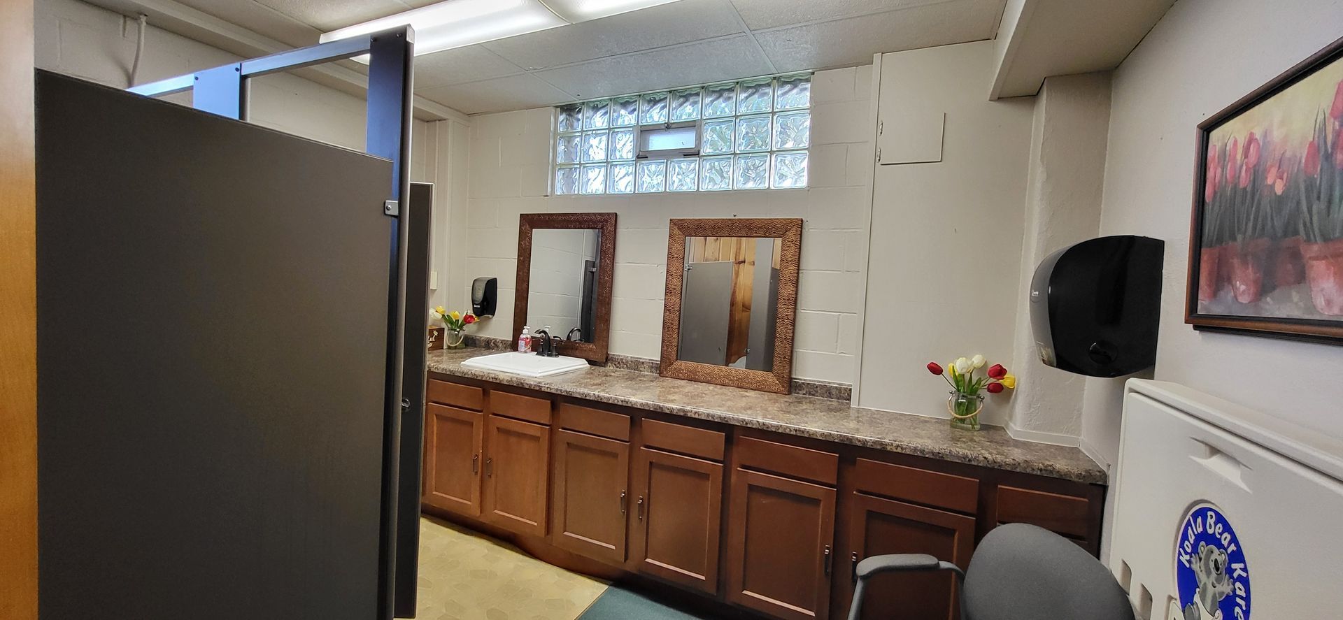 A public restroom with brown cabinets, two mirrors, and a window. A dark stall door is in the foreground.