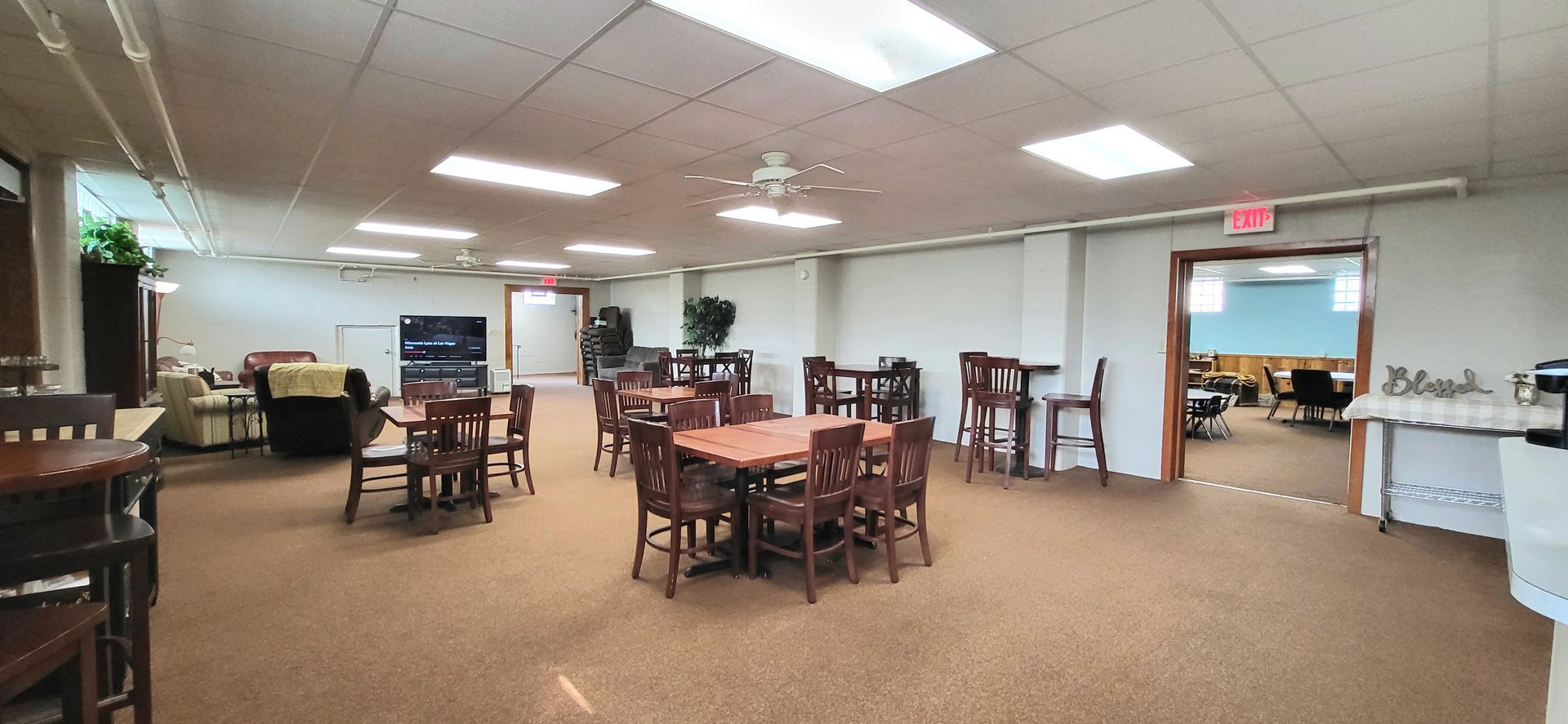 An interior shot of a large room, possibly a dining area or event space. There are tables, chairs, a sofa, and an open doorway.