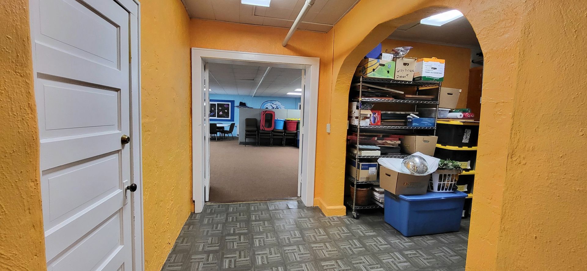 Hallway with yellow walls, a white door on the left, and an arched opening on the right with shelves and storage. A doorway leads to another room.