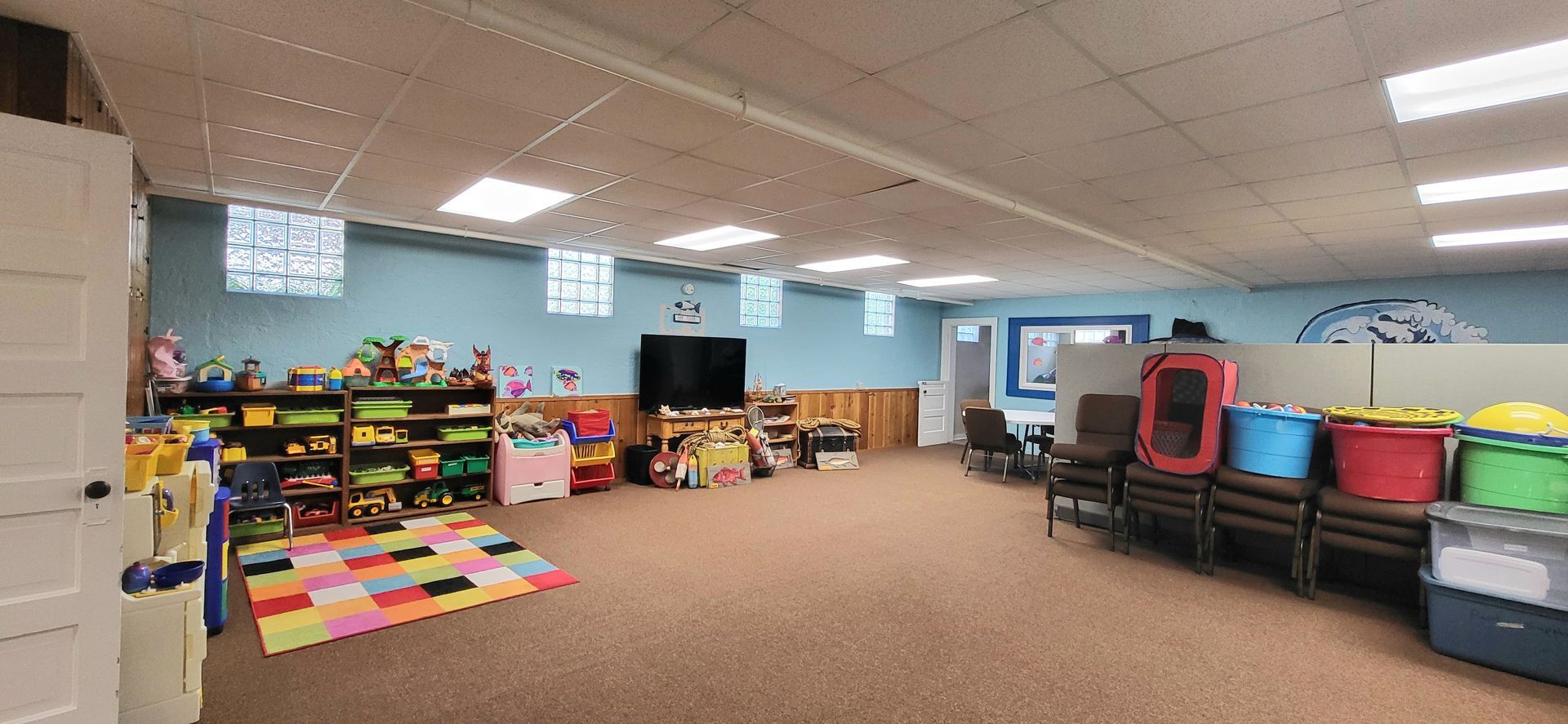 A playroom with toys, a colorful playmat, and a row of chairs. Blue walls, brown carpet, and bright overhead lights.
