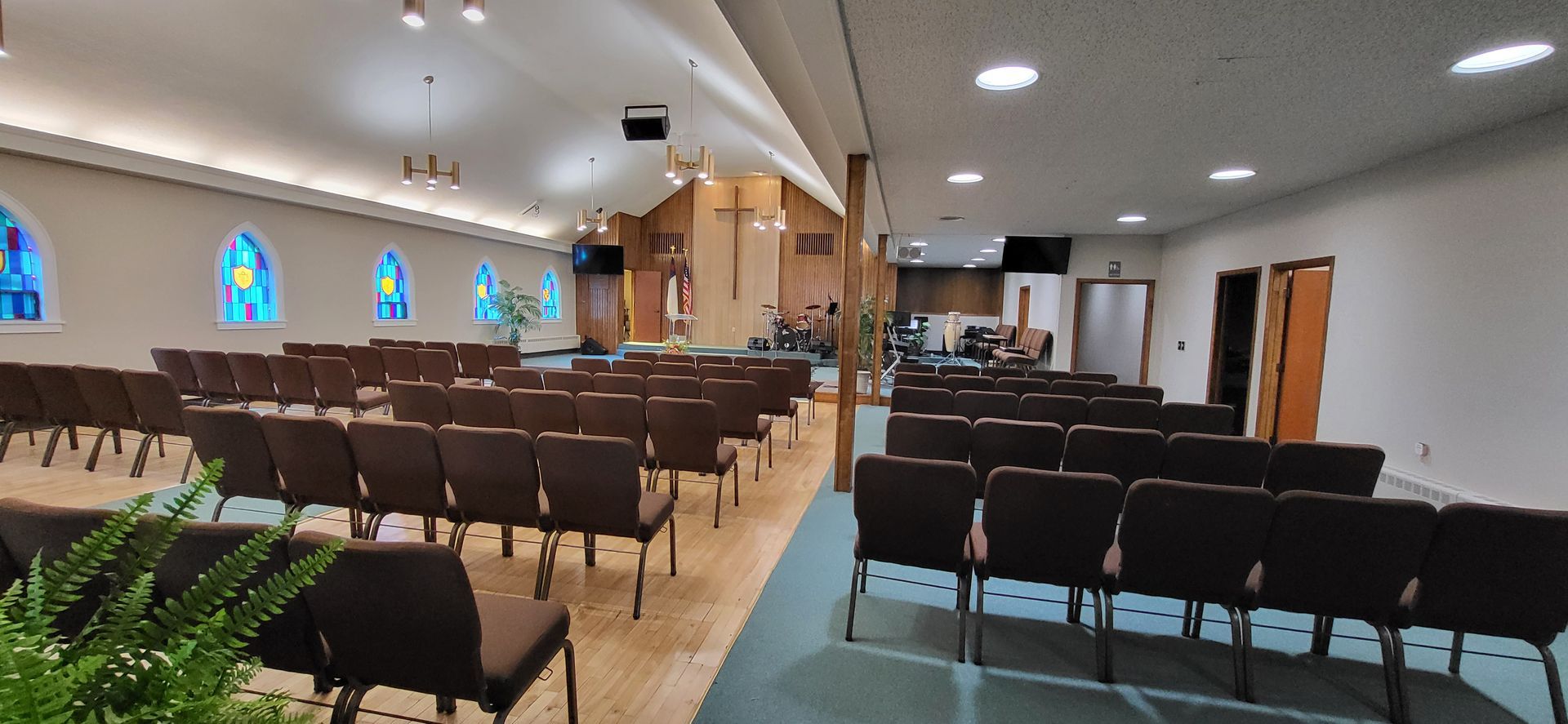 Interior of a church sanctuary with rows of brown chairs, stained glass windows, and a wooden cross on the stage.